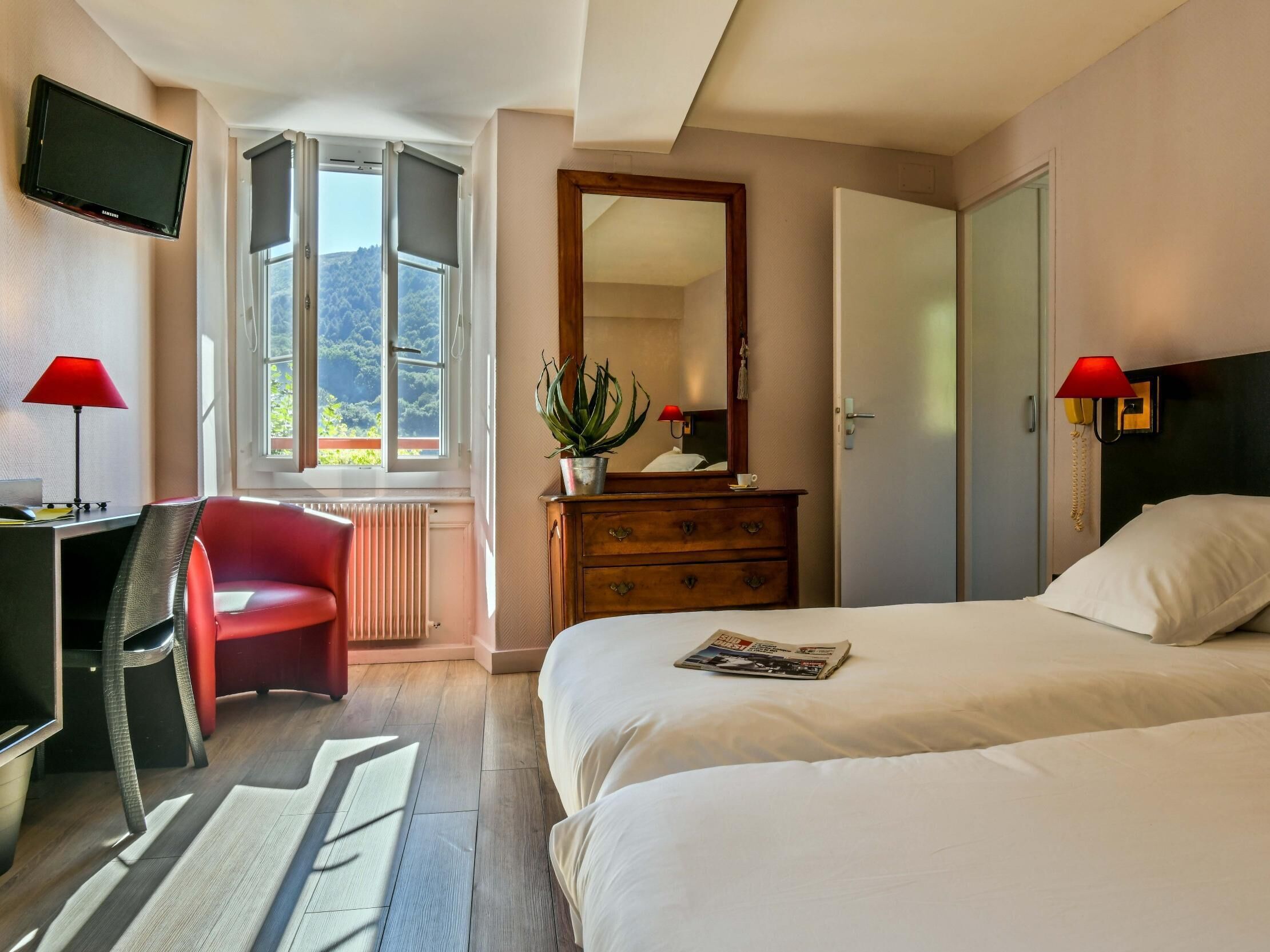 A hotel room with a bed, a desk with a chair, a TV, a dresser, a mirror, and a window with a view of trees.