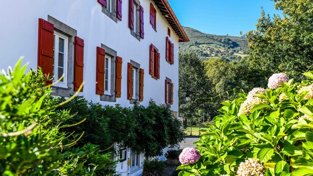 A white building with red shutters, surrounded by lush greenery and flowering plants.