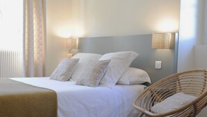 A neatly made bed with white linens and two decorative pillows, a wicker chair with a cushion, and wall-mounted lights on either side of the bed.