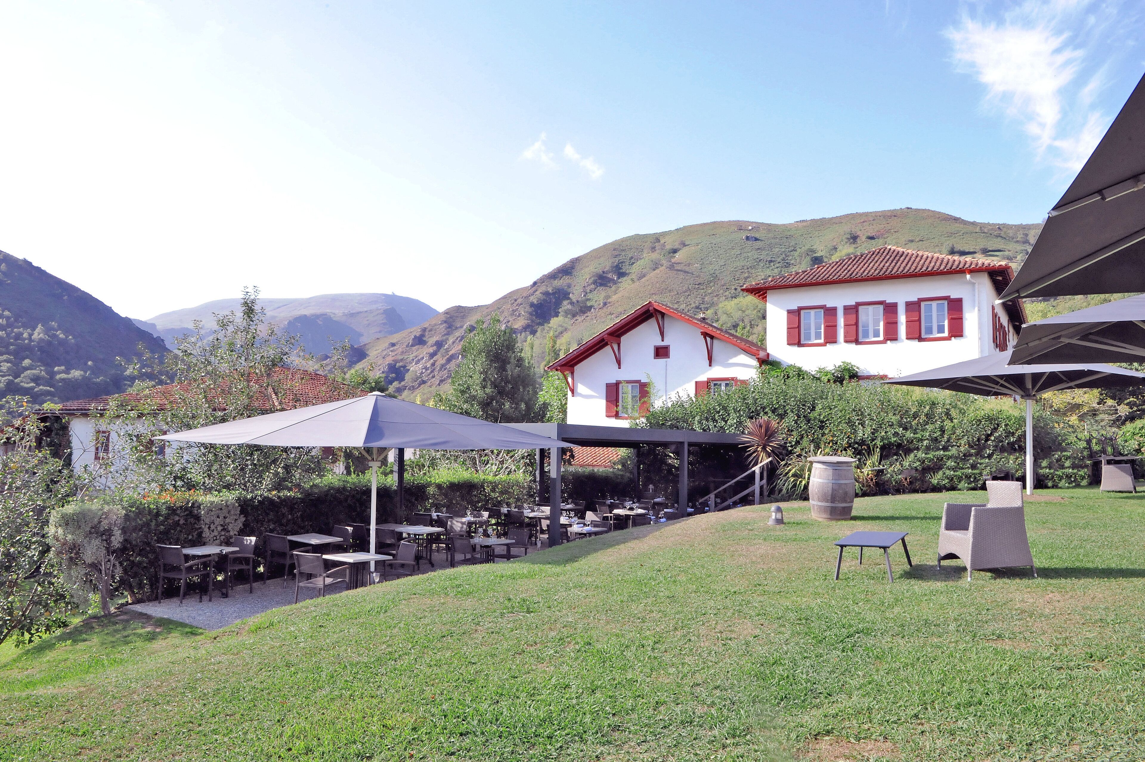 A patio area with outdoor seating, a white building with red shutters, and a mountainous backdrop.