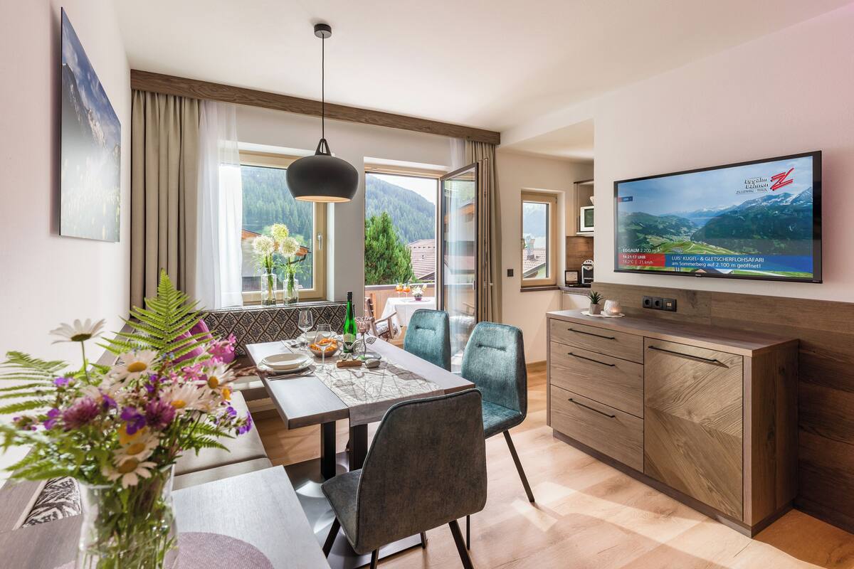 A modern dining area with a table set for two, a television mounted on the wall, and a view of mountains through the window.