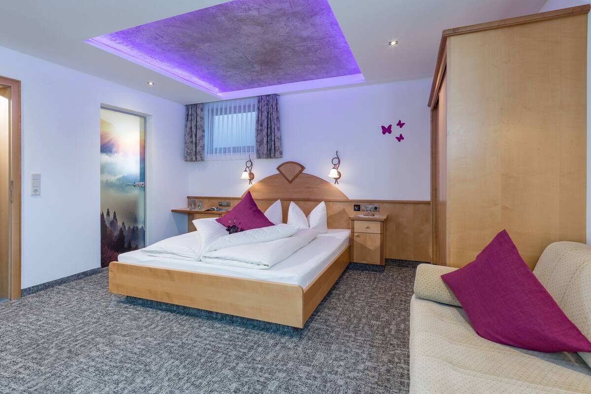 A modern hotel room with a large bed, a wooden headboard, a bedside table with a lamp, and a wardrobe.