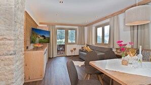 Una sala de estar moderna con pared de piedra, mobiliario de madera, televisor y comedor con mesa preparada para dos.