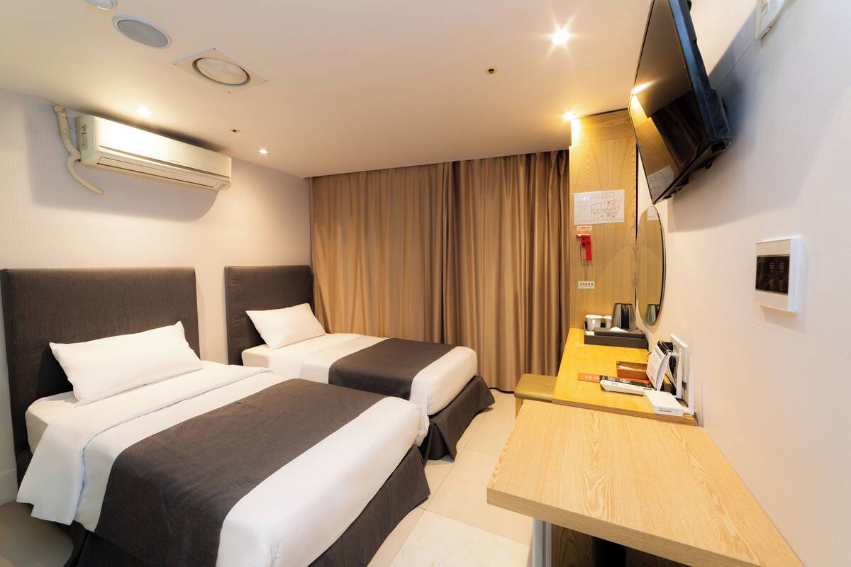 A hotel room with two beds, a desk, a TV, and an air conditioning unit.