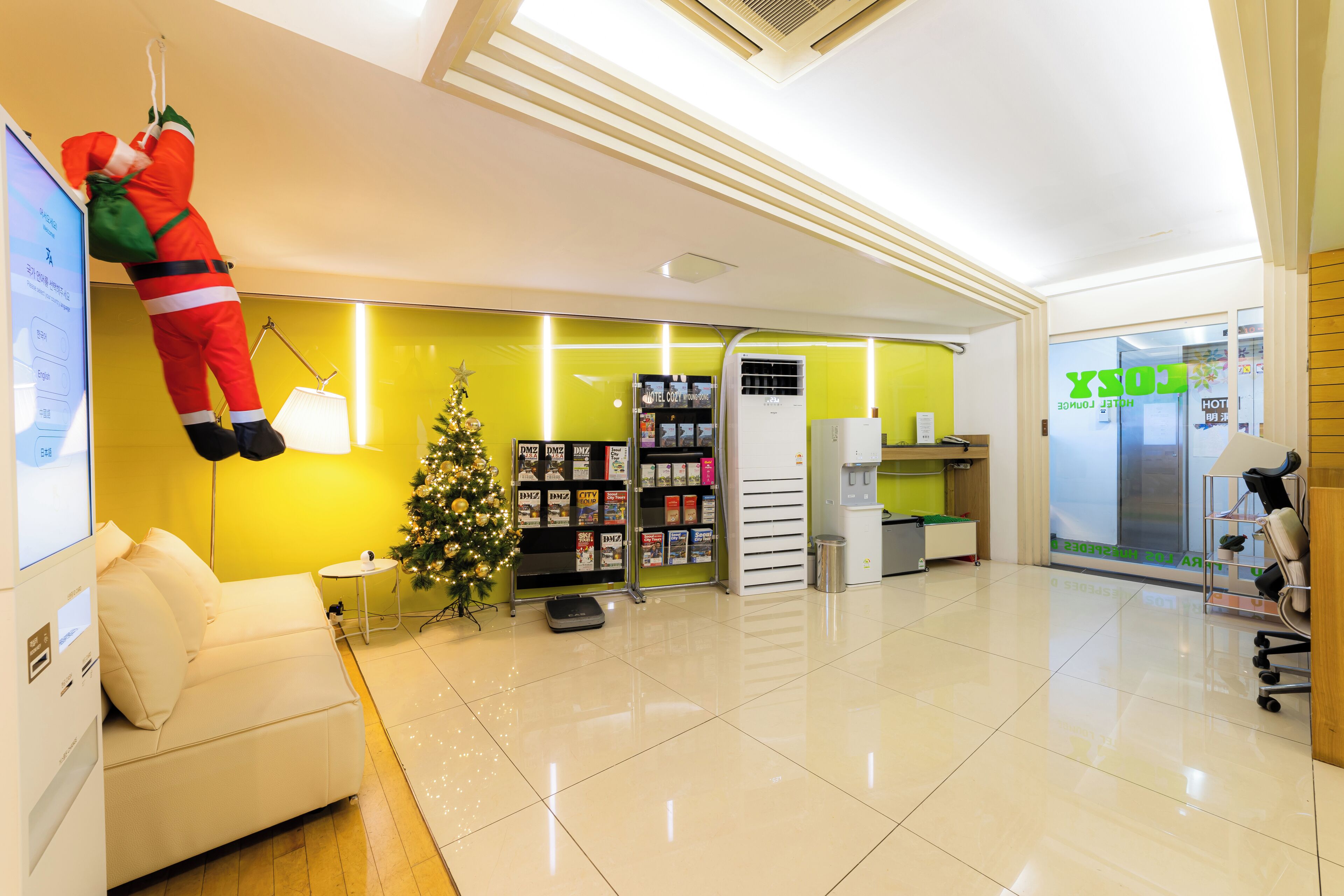 A hotel reception area with a white sofa, a Christmas tree, a hanging Santa figure, a reception desk, and a glass door with a sign.