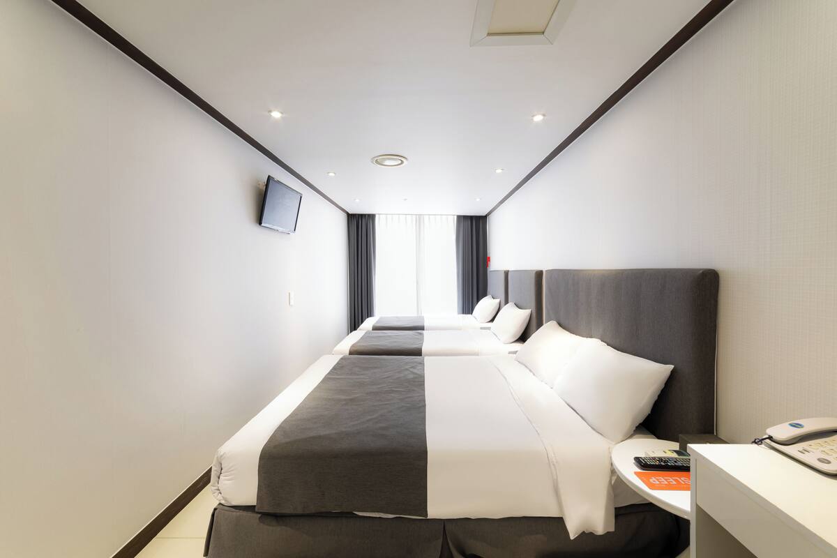 A hotel room with two beds, a TV mounted on the wall, a bedside table with a phone, and a view of the city through large windows.