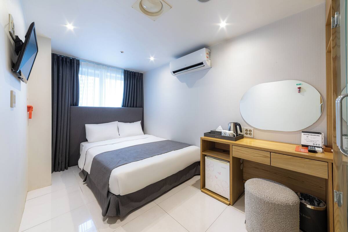 A hotel room with a bed, a desk, a chair, a mirror, a TV, and an air conditioning unit.