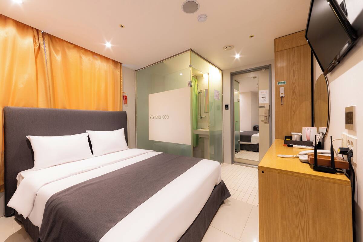 A hotel room with a bed, a desk, a television, and a glass partition.