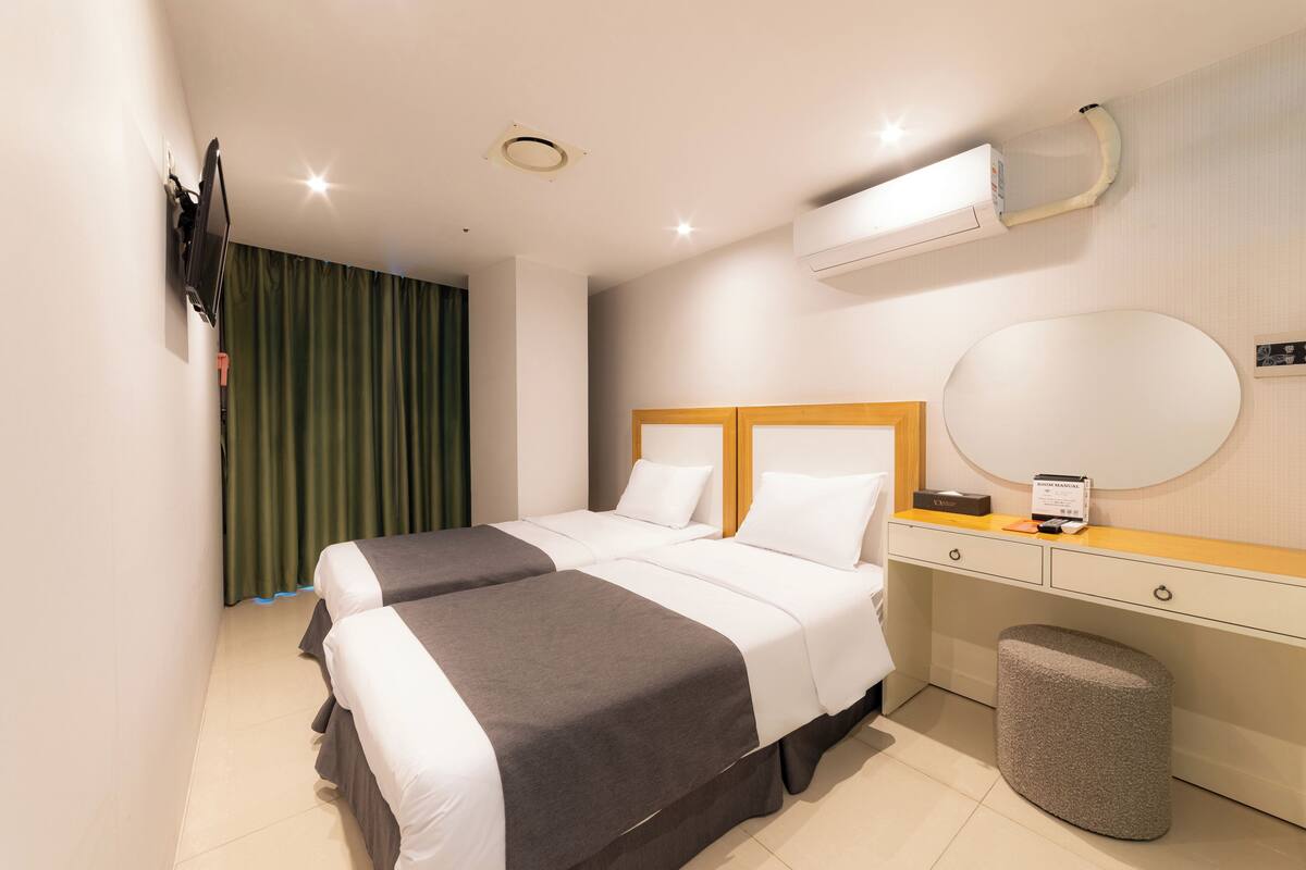 A hotel room with two beds, a wooden headboard, a bedside table with a lamp, a round mirror, and a waste bin.