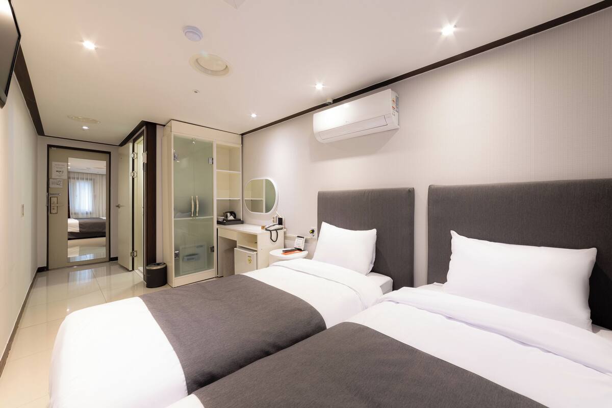 A modern hotel room with two beds, a desk, a mirror, and an air conditioning unit.