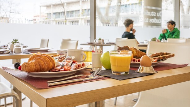 Een ontbijtsetting met croissants, fruit en drankjes op een tafel in een lichte kamer met grote ramen.