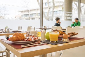 Une table dressée pour le petit-déjeuner, avec des croissants, des fruits et des boissons, dans une pièce lumineuse dotée de grandes fenêtres.