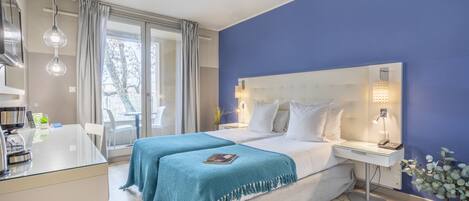Habitación de hotel moderna con una cama grande, una mesita de noche con lámpara, un escritorio pequeño con teléfono, una mesa de vidrio con cafetera y vistas al exterior a través de amplios ventanales.