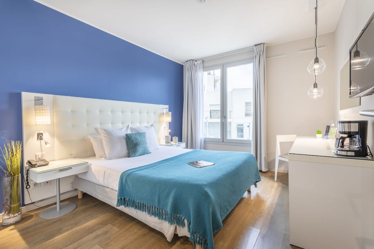 Een moderne hotelkamer met een groot bed, een nachtkastje met een lamp, een klein bureau met een koffiezetapparaat en uitzicht op de stad door het raam.
