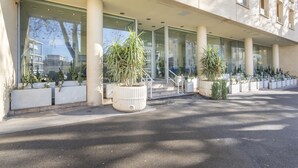 Een modern gebouw met grote ramen en een overdekte entree. Er staan verschillende potplanten langs het pad.