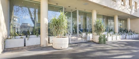 A modern building with large windows and a covered entrance area. There are several potted plants along the walkway.