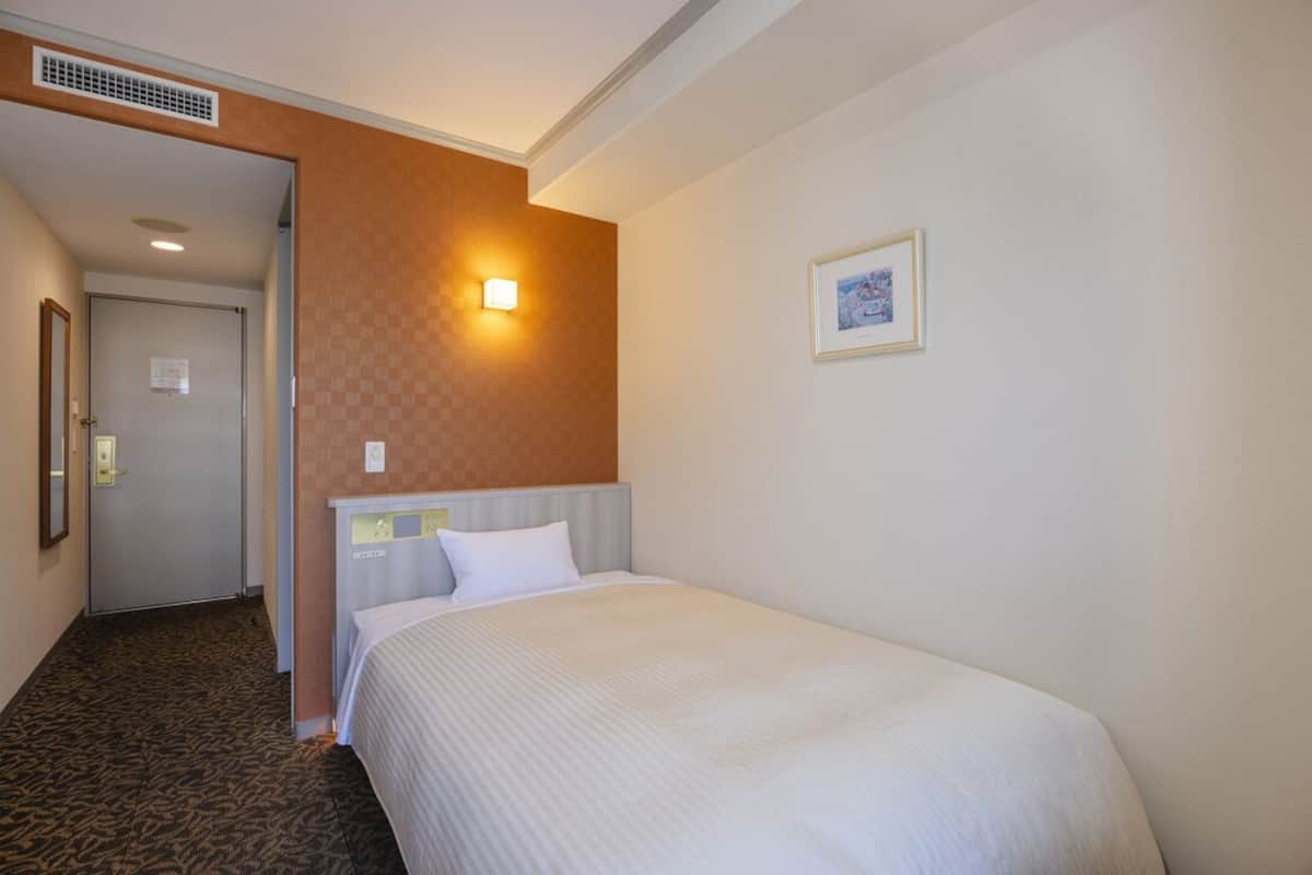 Habitación de hotel con cama, luz empotrada en la pared, un cuadro enmarcado y una puerta que da a otra habitación.