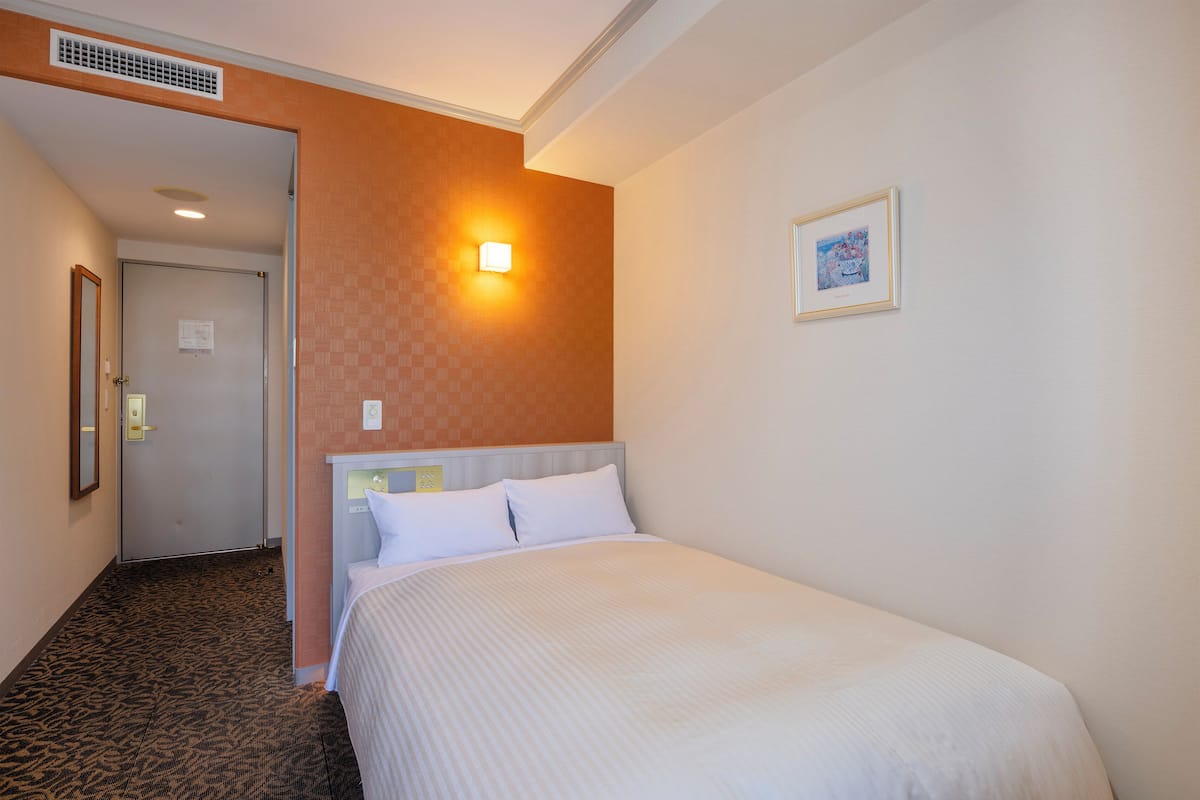 Una habitación de hotel con una cama, una mesita de noche, una lámpara de pared, un cuadro enmarcado y una puerta que da a otra habitación.
