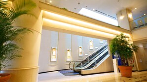 A modern hotel lobby with an escalator, potted plants, and a tiled floor.