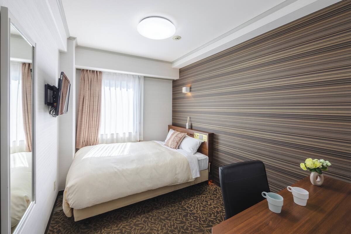 Una habitación de hotel con cama, escritorio con silla, una mesita con un florero y televisor montado en la pared.