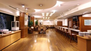 A spacious hotel dining area with wooden flooring, multiple tables and chairs, a buffet counter, and a television on the wall.