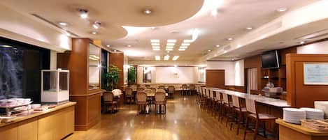 Ampla área de jantar de hotel com piso de madeira, várias mesas e cadeiras, um balcão de buffet e uma televisão na parede.