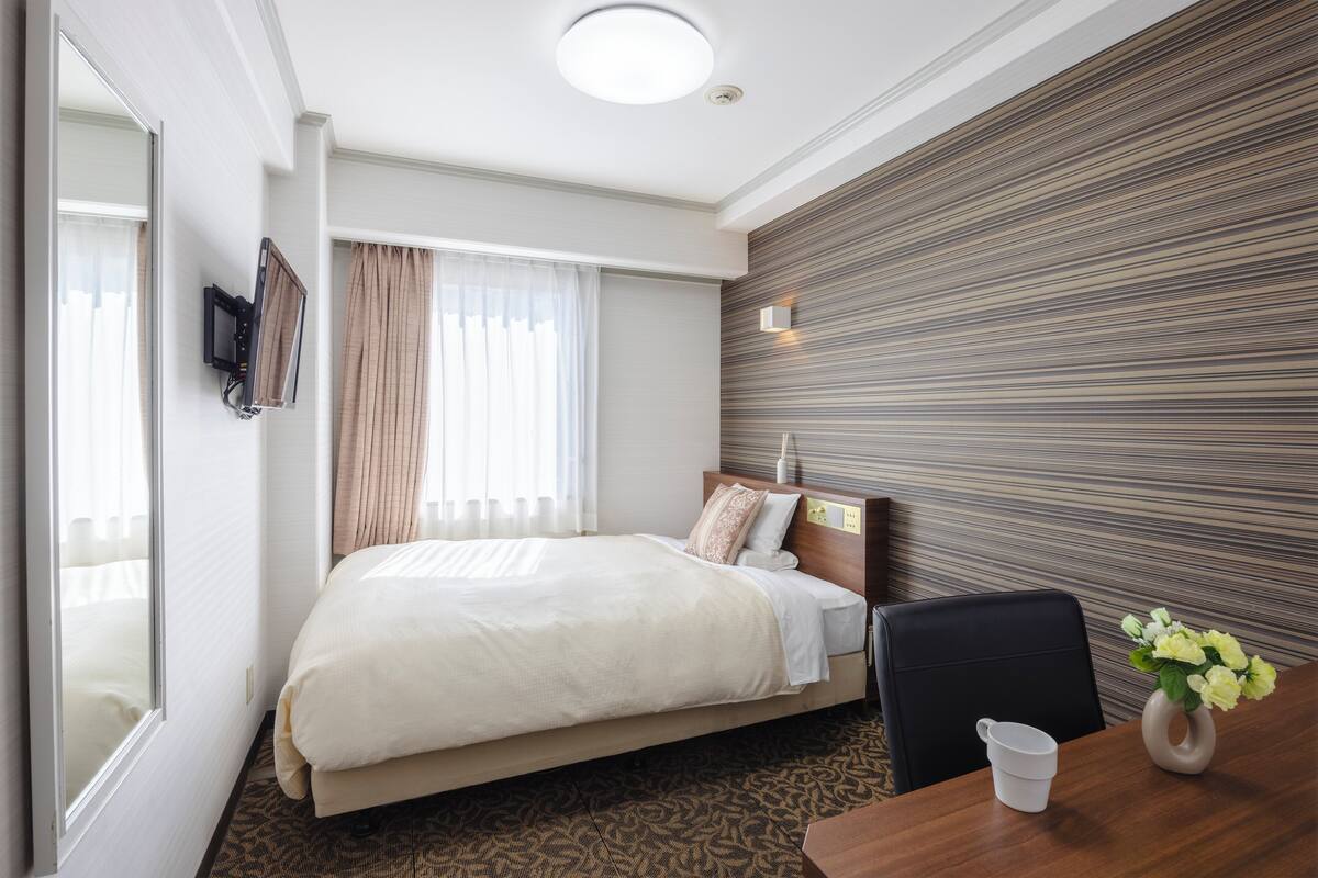 Habitación de hotel con cama, escritorio con silla, televisor montado en la pared y ventana con cortinas.