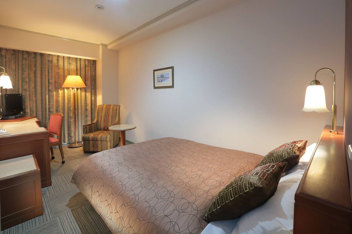 Habitación de hotel con cama, escritorio con computadora, silla, lámpara y un cuadro en la pared.
