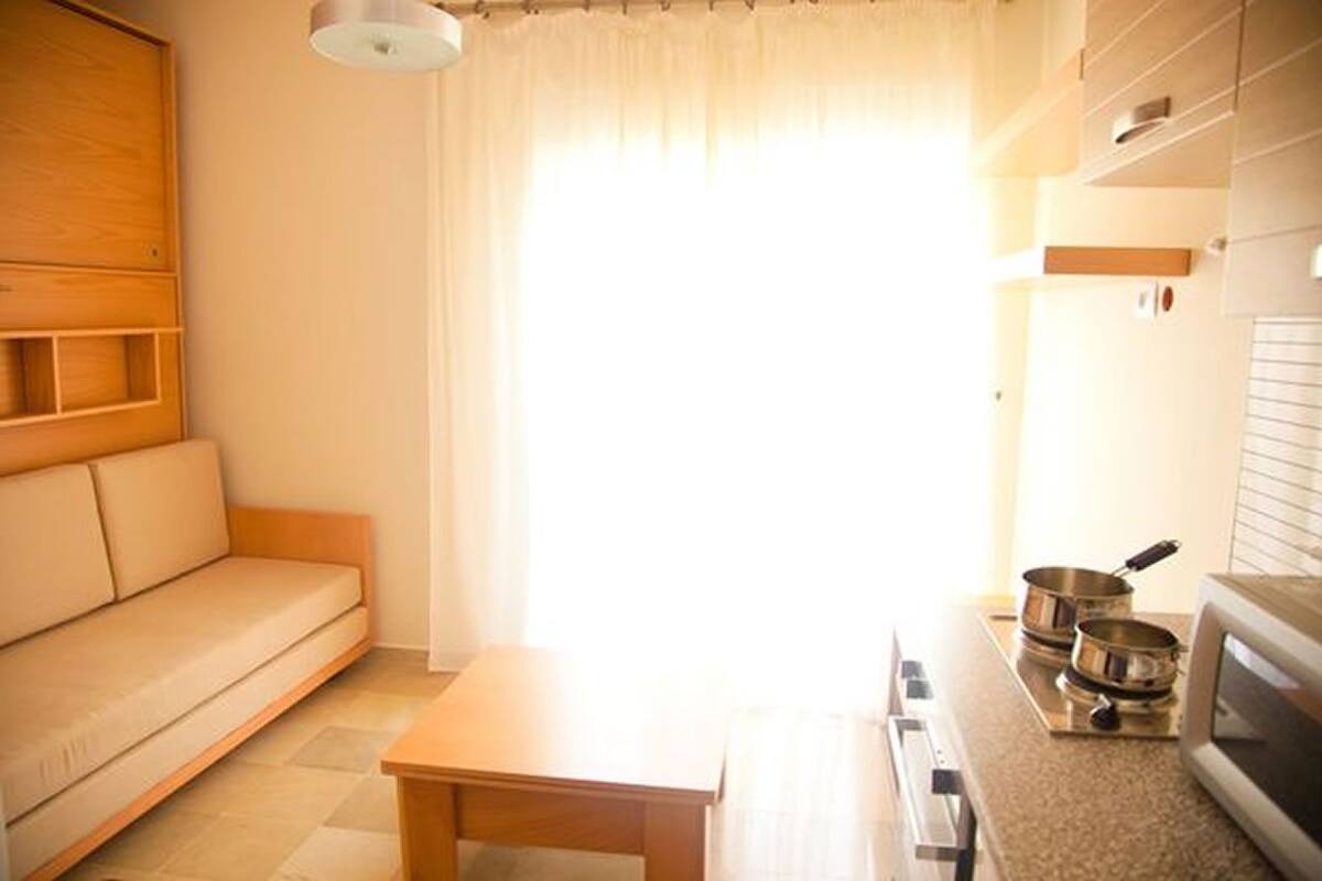 Un espace de vie compact comprenant un canapé, une petite table, une kitchenette équipée d’un four à micro-ondes et une fenêtre agrémentée de rideaux légers.