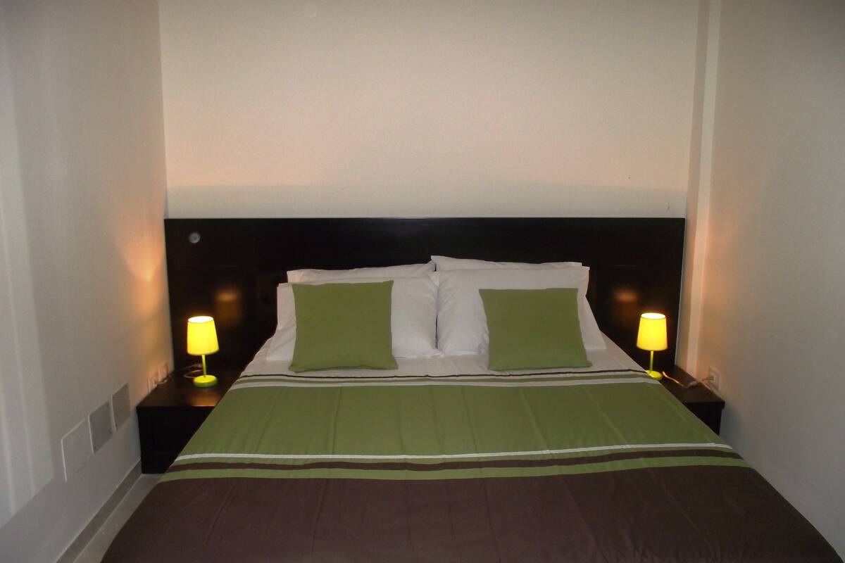 Un lit bien fait, recouvert d’une couvre-lit verte et marron, avec deux oreillers verts et deux lampes de chevet dotées d’abat-jour jaunes.