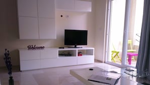 A modern living room with a flat-screen TV on a white entertainment unit, a glass coffee table with a laptop, and a view of outdoor furniture through a glass door. - The Dunes Golf & Spa Resort (Port El Kantaoui)