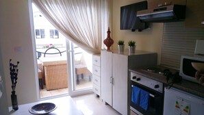A kitchen with a microwave, oven, and cabinets. There is a window with curtains, a wicker chair outside, and a small table with a bowl. - The Dunes Golf & Spa Resort (Port El Kantaoui)