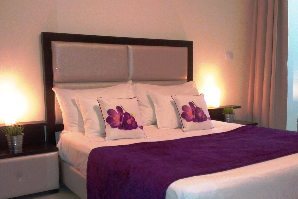 Un lit bien fait, agrémenté d’un plaid violet, de draps blancs et de deux coussins décoratifs ornés de fleurs violettes. On y trouve deux tables de chevet avec des lampes, et une plante en pot sur l’une d’elles.