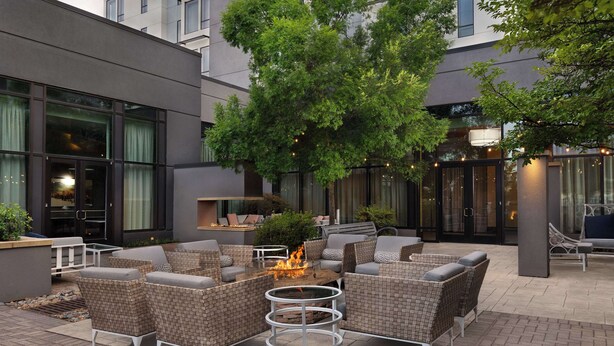 Un patio al aire libre con mobiliario de mimbre, una chimenea central y un edificio moderno al fondo.