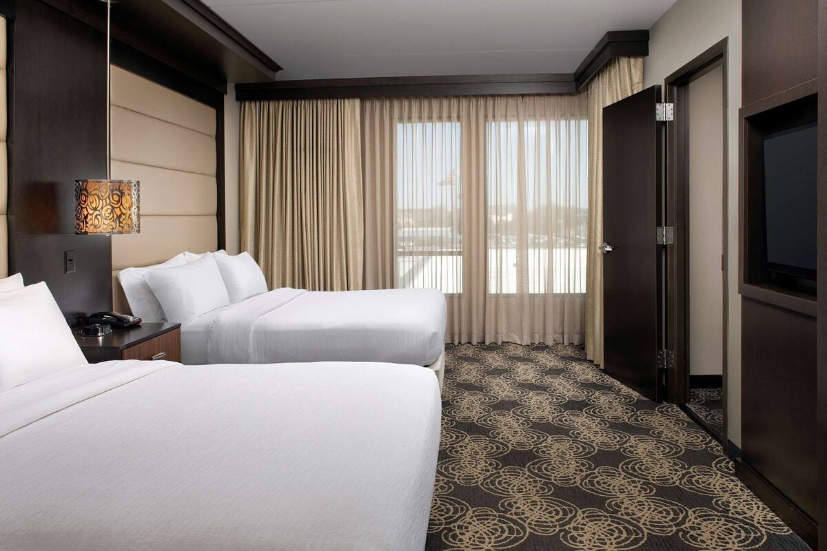 A hotel room with two beds, a desk with a lamp, a television, and a large window with curtains.