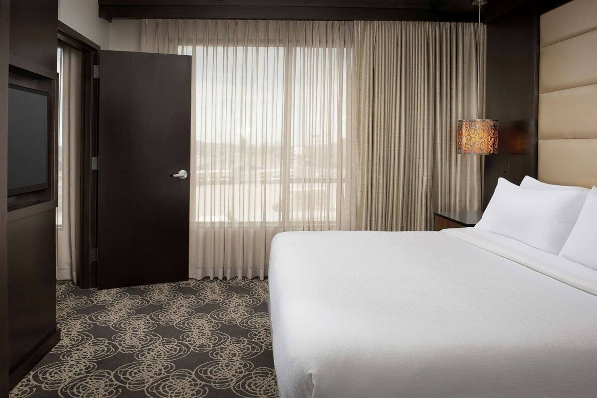 A hotel room with a bed, a door, a window with sheer curtains, a patterned carpet, and a wall-mounted lamp.