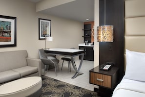 A hotel room with a sofa, a desk, a chair, a bed, a nightstand, a lamp, and a phone.