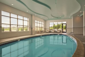An indoor swimming pool with clear blue water, surrounded by lounge chairs and a view of greenery outside.