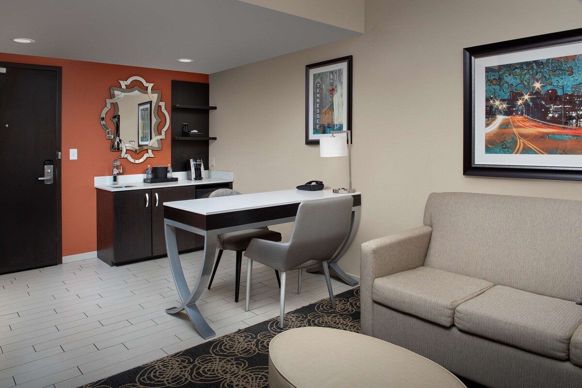 A hotel room with a desk, chair, sofa, and a framed cityscape picture on the wall.