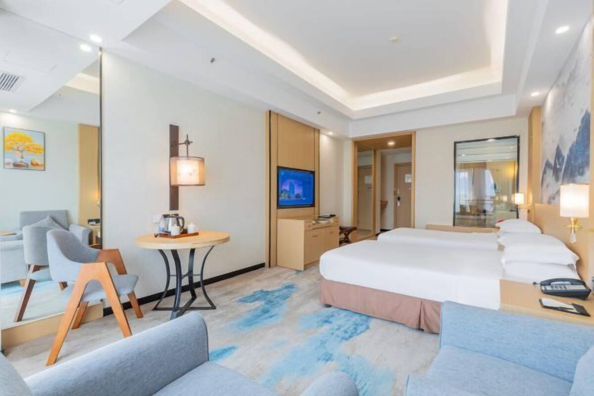 A hotel room with a bed, a desk, a chair, a table with a lamp, and a TV.