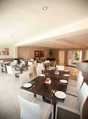 A spacious dining area with multiple tables and chairs, a central island with a water feature, and a view of the outdoor garden. - The Capital Villa (Sandton)