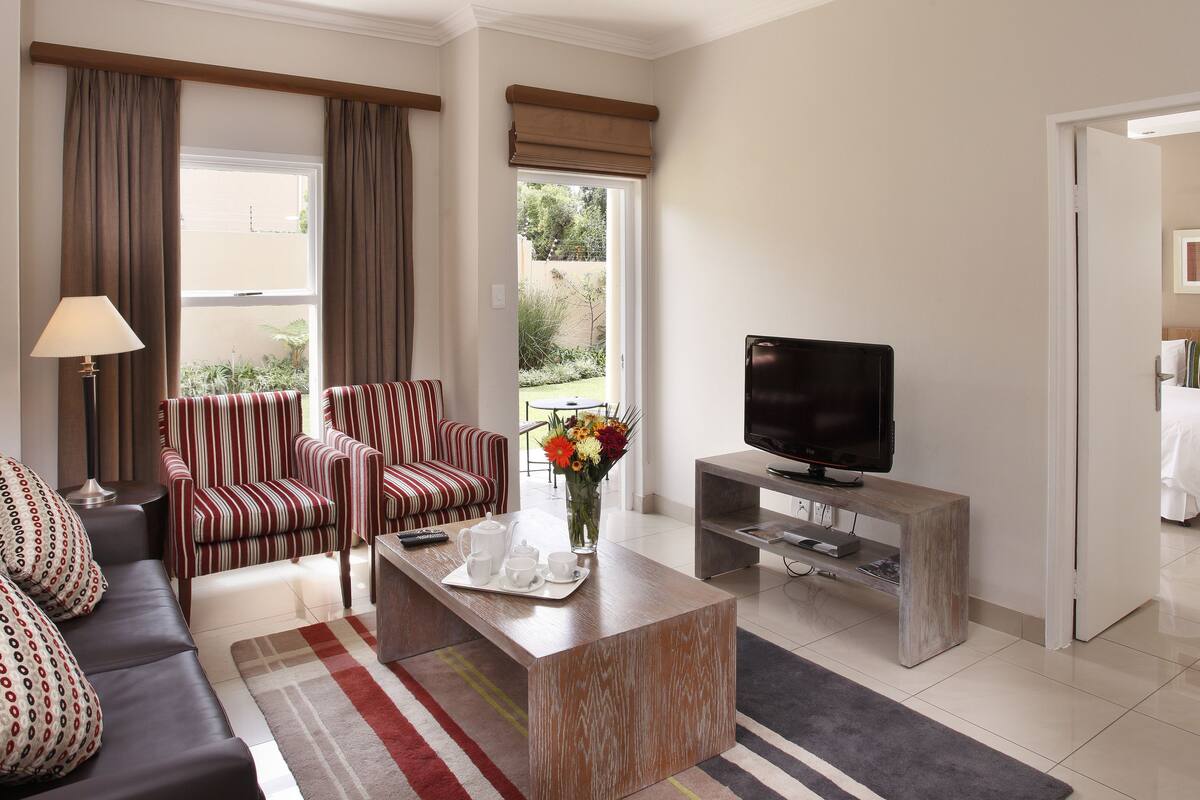A modern living room with a sofa, striped armchairs, a coffee table, a TV on a wooden stand, and a view of a garden through a window.