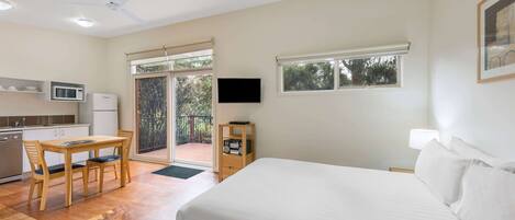 A hotel room with a bed, a desk with chairs, a television, and a balcony with a view of trees.