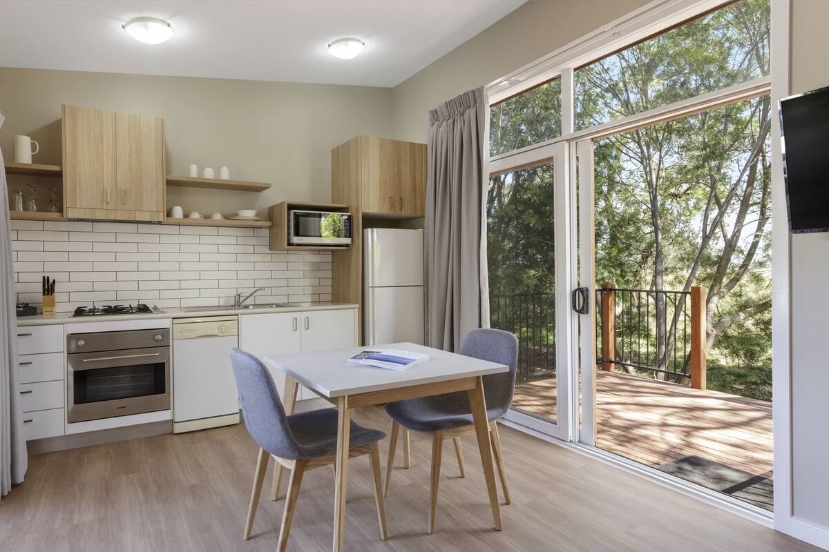 Eine moderne Küche mit Holzausstattung, einer Mikrowelle und einem Kühlschrank. Ein Esstisch mit zwei Stühlen bietet Blick auf eine Terrasse durch große Schiebeglastüren.