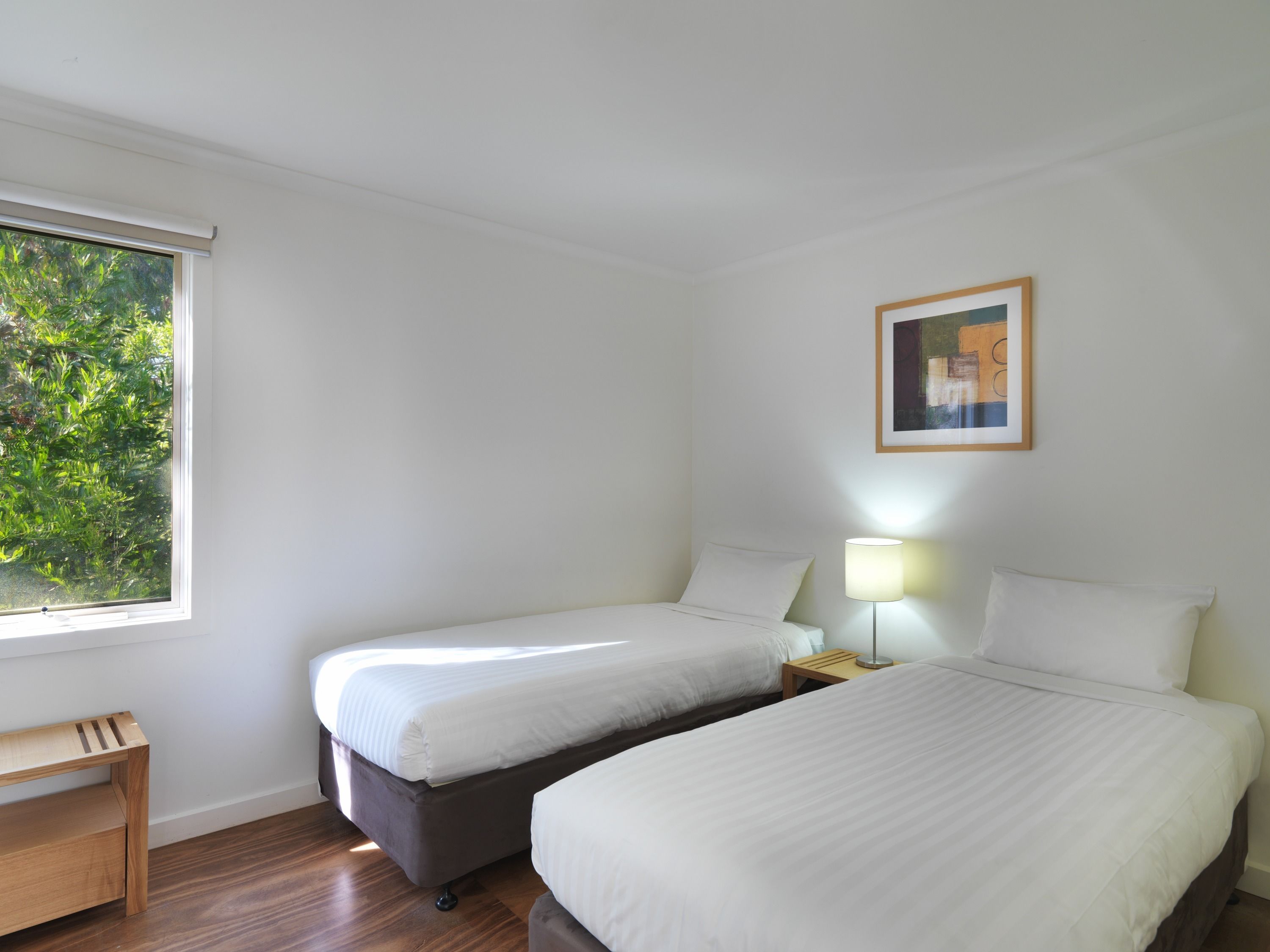 Habitación de hotel con dos camas, una mesita de madera, una mesita de noche con lámpara y ventana con vistas a zonas verdes.