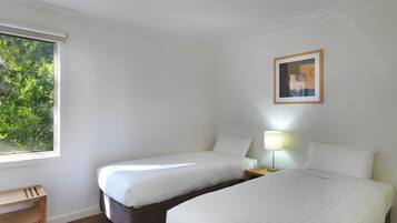 Habitación de hotel con dos camas, una mesita de madera, una mesita de noche con lámpara y ventana con vistas a zonas verdes.