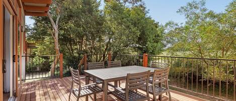Un terrazzo in legno con tavolo e sedie, circondato da una ringhiera in metallo, affacciato su un giardino con alberi e arbusti.