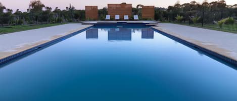 Ein rechteckiger Pool mit glasklar blauem Wasser, umgeben von Kiesflächen und einem Metallzaun. Im Hintergrund sind drei Holzbauten mit Lamellen-Dächern zu sehen.
