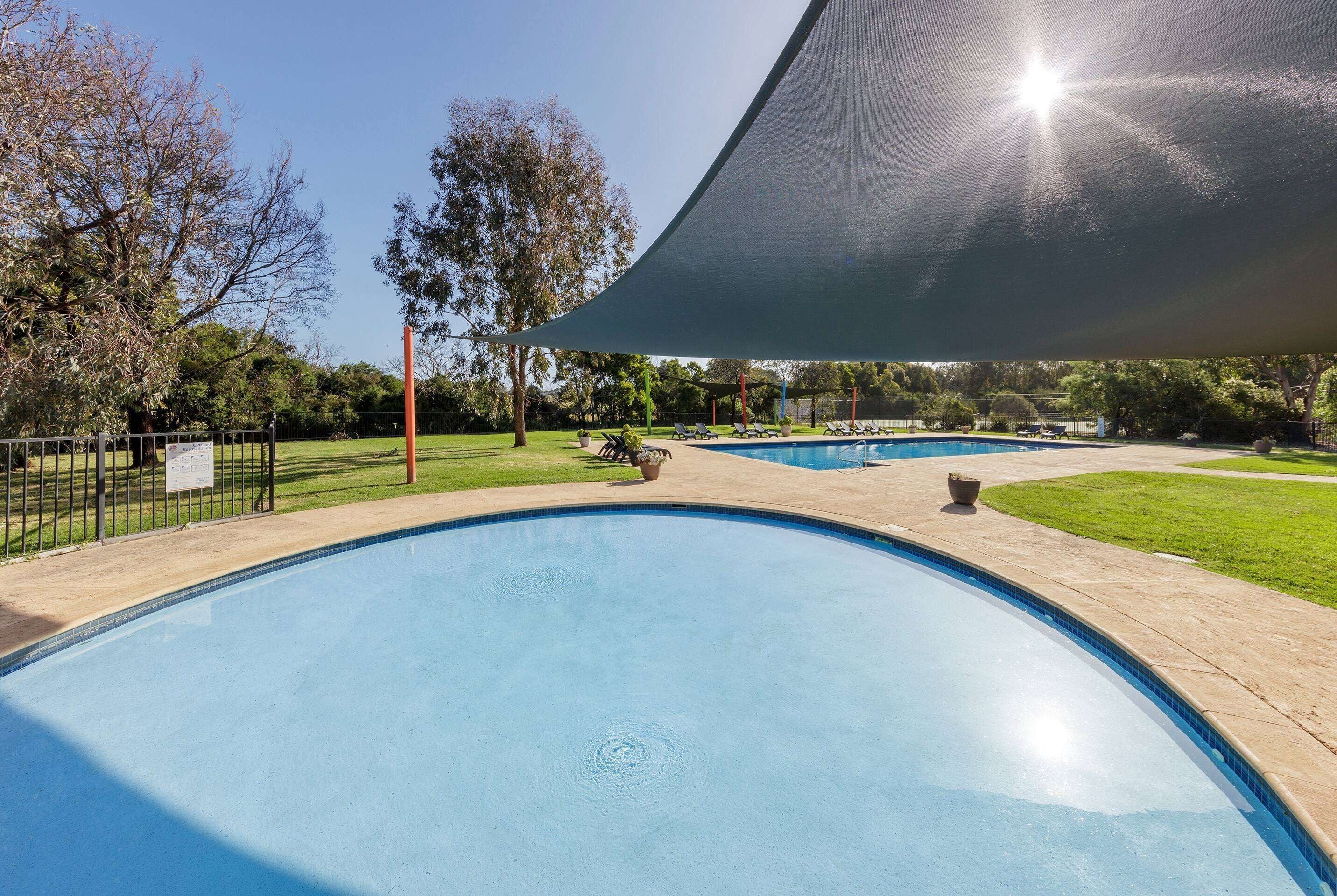 A swimming pool with a clear blue water surface, surrounded by a paved walkway and a grassy area with trees.