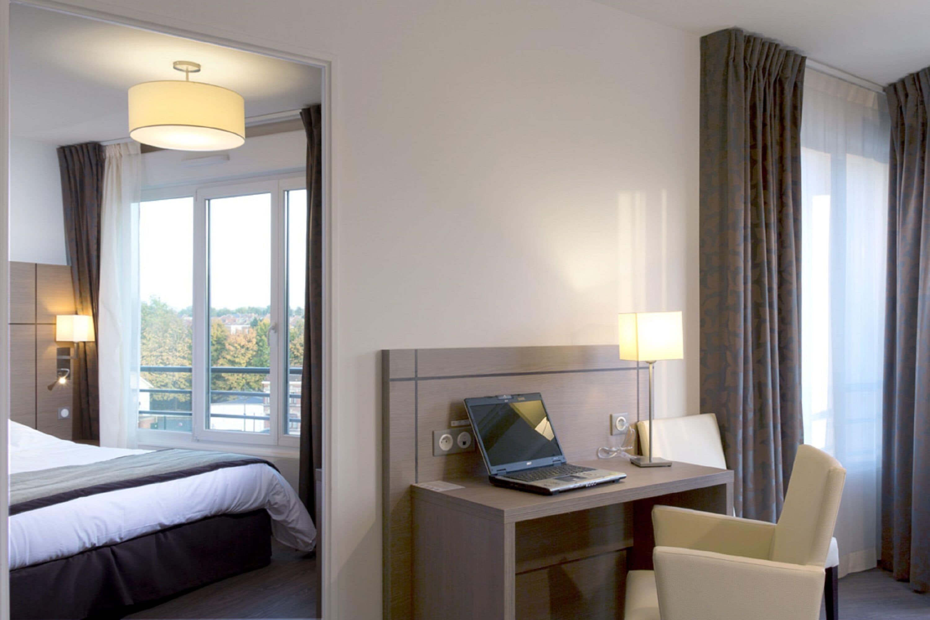 Quarto de hotel com cama, escrivaninha, um laptop, uma cadeira e janela com cortinas.